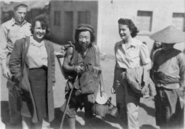 Eileen Denney and Betty Ringland stand on each side of a roving food vendor with walking stick in his right hand; all facing camera.  He holds buckets of rice and a plate in left hand, had has carry sacks with unidentifiable objects on his back and chest.